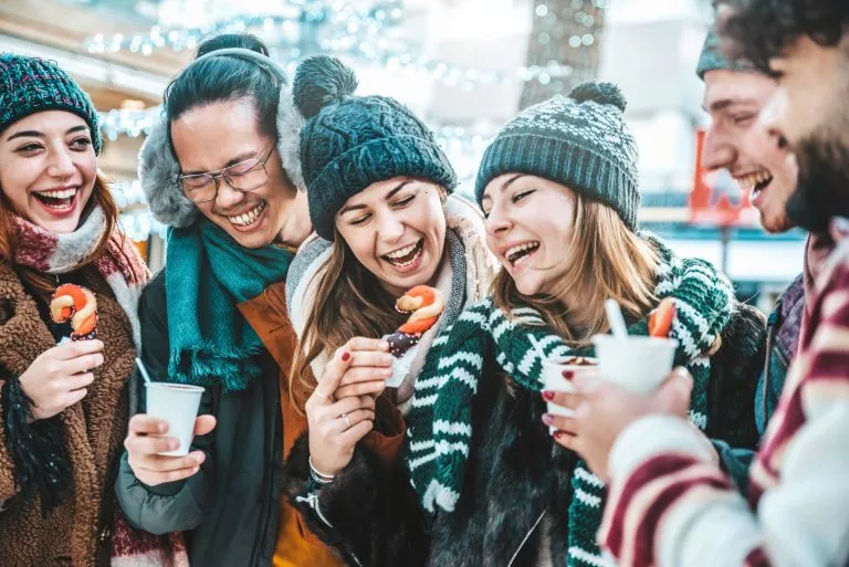 Happy friends having fun drinking mulled wine and hot chocolate at Christmas Market - Cheerful young people enjoying winter holidays on weekend vacation - Focus on asian guy