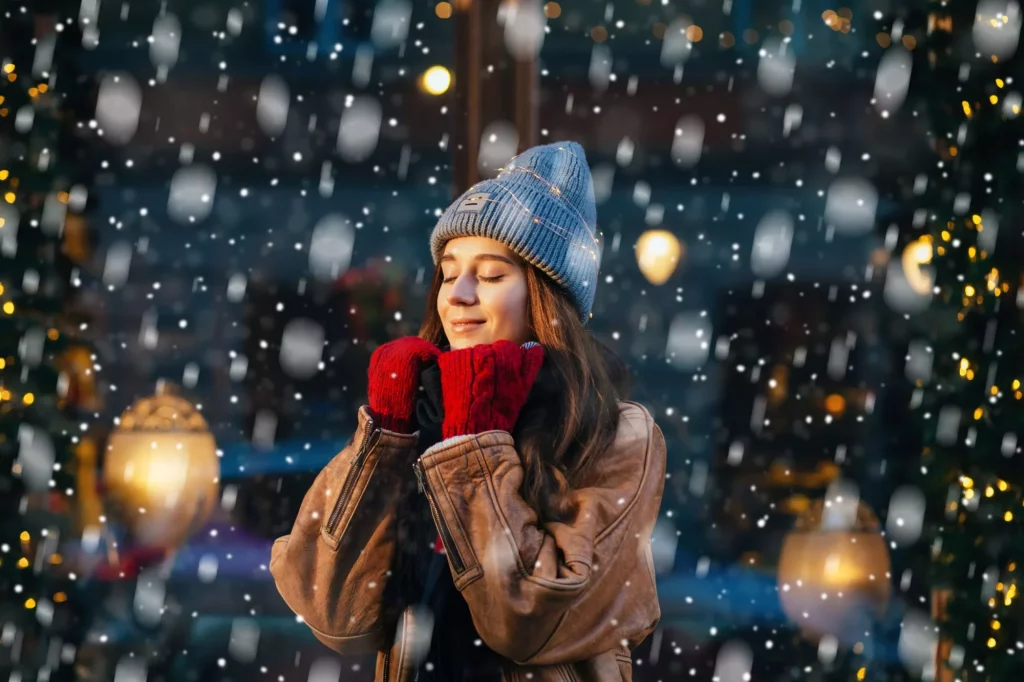 Cute woman closing eyes and dreaming in winter city a festive mood. Snowfall. Magic light. Christmas, new year, winter holidays concept.